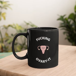 以下が含まれることがあります： 黒いセラミックマグカップ。白い文字で「FUCKING OVARY-IT」と、子宮と卵巣のピンク色のイラストが描かれています。マグカップには黒いハンドルがあり、木製の表面に置かれています。