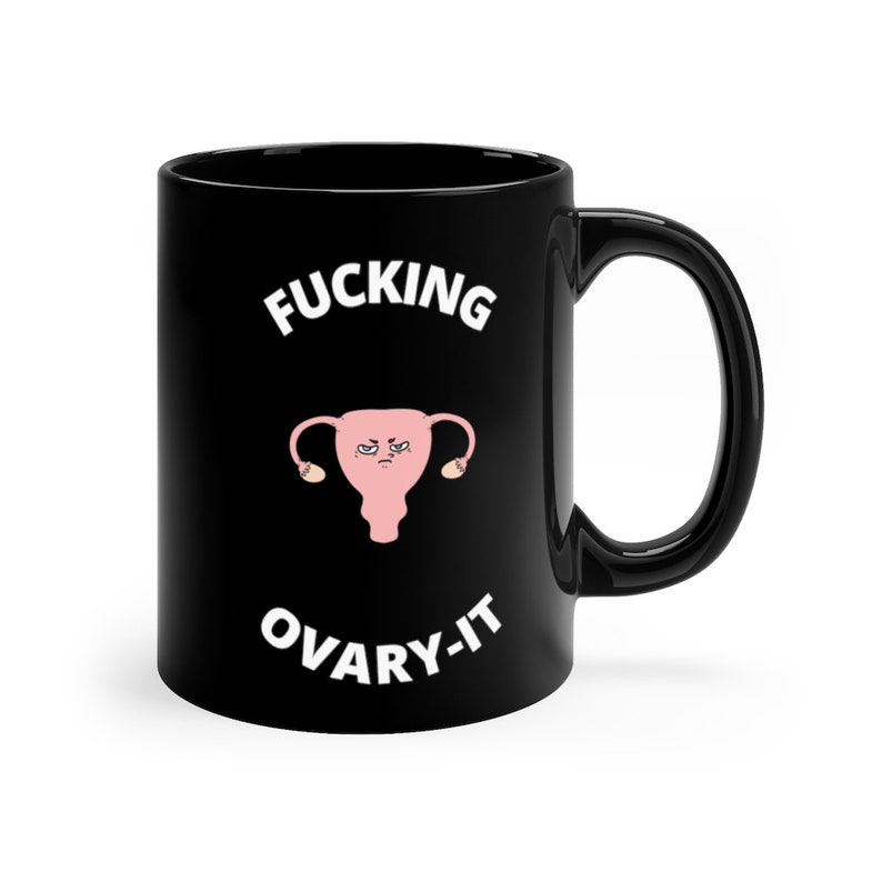 以下が含まれることがあります： 「FUCKING OVARY-IT」の白文字が入った黒いセラミックマグカップ。怒った顔の卵巣のピンク色のイラストが特徴です。マグカップは黒いハンドルと光沢のある仕上がりです。