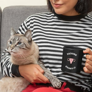 以下が含まれることがあります： 「FUCKING OVARY-IT」の文字とピンク色の子宮のグラフィックが描かれた黒いセラミックマグカップ。 黒と白のストライプの長袖シャツを着た人がマグカップを持っています。 猫がその人の膝の上に座っています。