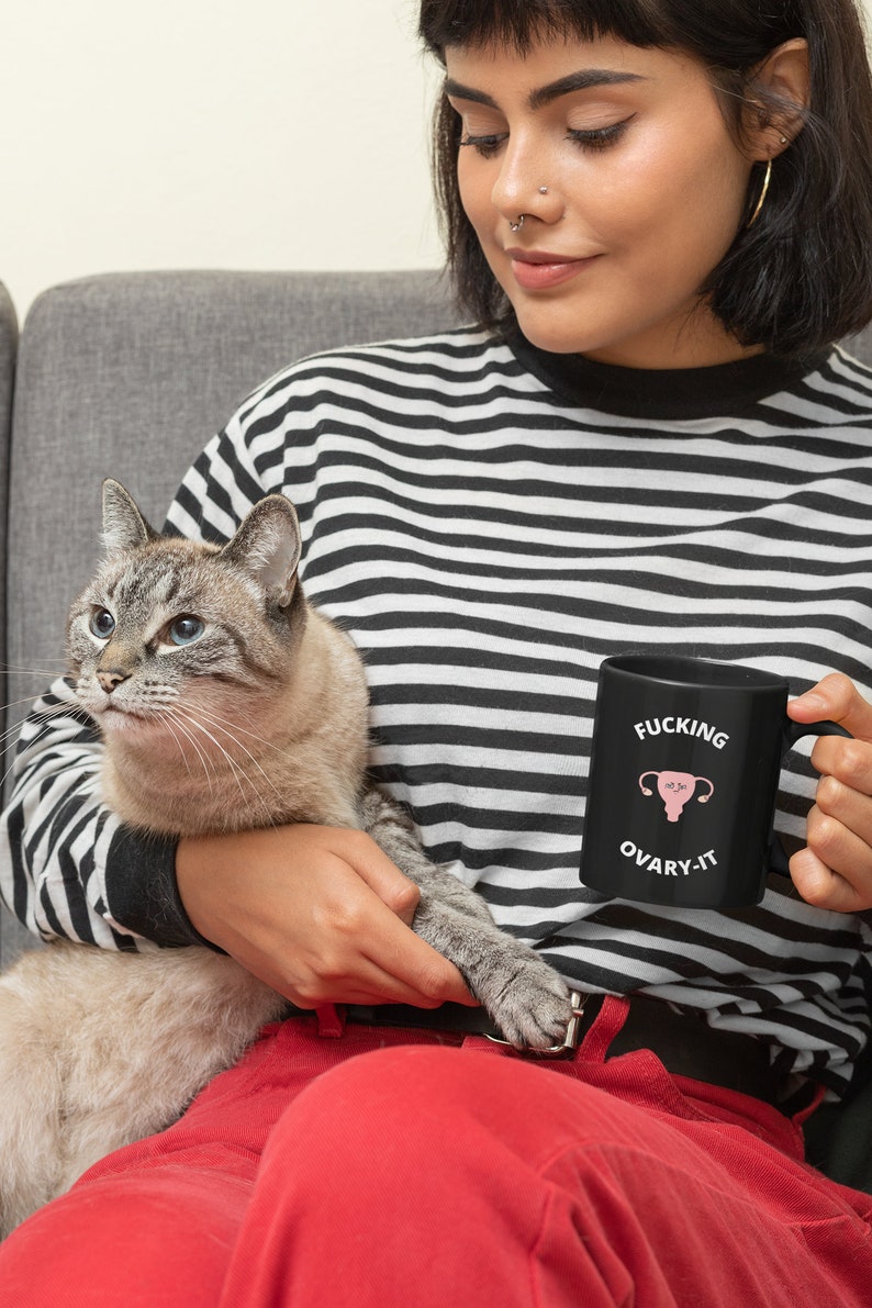 以下が含まれることがあります： 「FUCKING OVARY-IT」の文字とピンク色の子宮のグラフィックが描かれた黒いセラミックマグカップ。 黒と白のストライプの長袖シャツを着た人がマグカップを持っています。 猫がその人の膝の上に座っています。