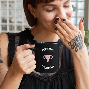 以下が含まれることがあります： 「FUCKING OVARY-IT」の文字とピンク色の子宮のイラストが描かれた黒いセラミックマグカップ。マグカップは黒い持ち手があり、黒いトップスを着た人が持っています。