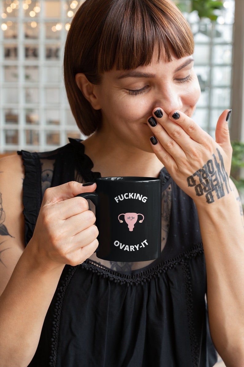 以下が含まれることがあります： 「FUCKING OVARY-IT」の文字とピンク色の子宮のイラストが描かれた黒いセラミックマグカップ。マグカップは黒い持ち手があり、黒いトップスを着た人が持っています。