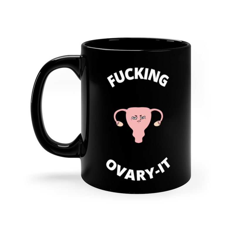 以下が含まれることがあります： 黒いセラミックマグカップ。白い文字で「FUCKING OVARY-IT」と書かれています。マグカップには、怒った表情のピンク色の漫画の子宮が描かれています。マグカップには黒いハンドルと白い背景があります。