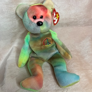 May include: A colorful tie-dye teddy bear plush toy with a peace sign on its chest. The bear has a red tag attached to its ear. The bear's fur is a mix of rainbow colors, including pink, green, blue, and yellow.