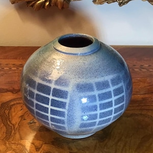 May include: A round, blue and white ceramic vase with a grid pattern. The vase has a glazed finish and a slightly rounded opening.