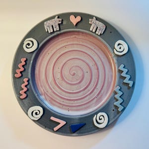 May include: A round ceramic plate with a dark gray rim and a light pink center featuring a spiral pattern. The rim is decorated with applied clay shapes including white spirals, pink and blue zigzags, two speckled animal figures, and a pink heart.