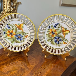 May include: Two round ceramic plates with openwork lattice rims and yellow painted edges. The center of each plate features a hand-painted floral motif with orange, blue, and green flowers and leaves on a white background, displayed on gold stands.