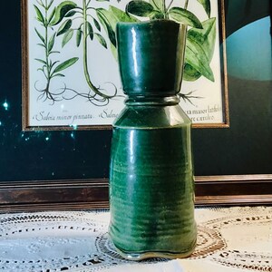 May include: A tall, cylindrical ceramic vessel with a glossy, deep forest green glaze. The object features a distinct horizontal ridge separating the narrower upper section from the wider, slightly tapered base, showcasing a smooth, reflective finish.
