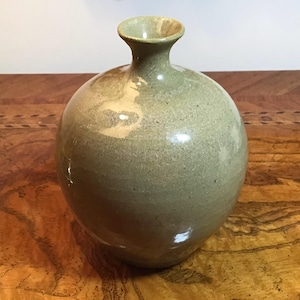 May include: A spherical ceramic vase with a narrow, flared neck and a smooth, glossy finish. The vase features a muted sage green color with subtle variations in tone and light-colored highlights across the rounded body and the rim.