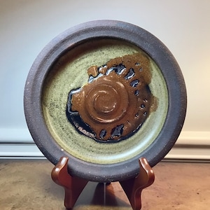 May include: A brown ceramic plate with a raised rim and a green and brown glaze. The plate has a unique design in the center, featuring a brown swirl with black accents.