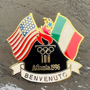 Può includere: Una spilla in metallo dorato con la bandiera americana e la bandiera italiana ai lati di uno scudo nero centrale. Lo scudo mostra i cerchi olimpici, il numero 100 e la scritta Atlanta 1996. Un nastro bianco in basso riporta la scritta BENVENUTO.