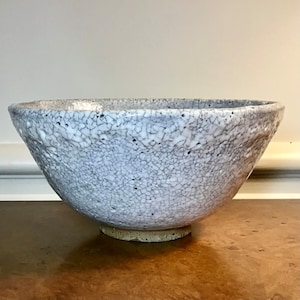 May include: A ceramic bowl with a textured, crackled white and grey glaze finish. The exterior surface features a dense network of fine dark fissures across the entire body, with a thick, uneven rim and a circular, unglazed tan base at the bottom.