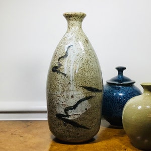 May include: A tall, speckled ceramic vase with a narrow neck and a rounded body. The vase features black abstract designs and a light brown and beige color scheme. Two other ceramic vessels are in the background.