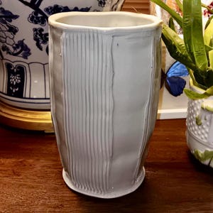 Puede incluir: Un jarrón de cerámica blanco con un diseño texturizado y acanalado. El jarrón tiene una forma ligeramente redondeada y una abertura ancha.