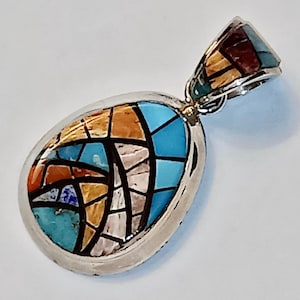 May include: A silver pendant featuring a turquoise, orange, and white inlay design. The pendant is oval shaped and has a bail for attaching a chain.
