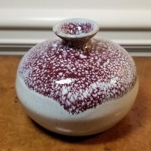 Puede incluir: Un pequeño jarrón redondo de cerámica con un esmalte blanco y un patrón de salpicaduras burdeos. El jarrón tiene un cuello estrecho y una base ancha y redondeada.