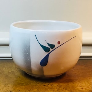 May include: White ceramic bowl with a blue, green, and red abstract design. The bowl has a slightly rounded shape and a smooth, glossy finish.
