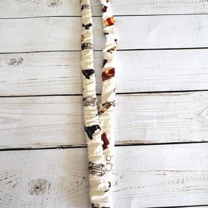 May include: A white fabric lanyard with a repeating pattern of black silhouettes, gold accents, and red and gold crests. The lanyard has a metal clasp and ring at the bottom. The fabric is gathered, creating a textured appearance.