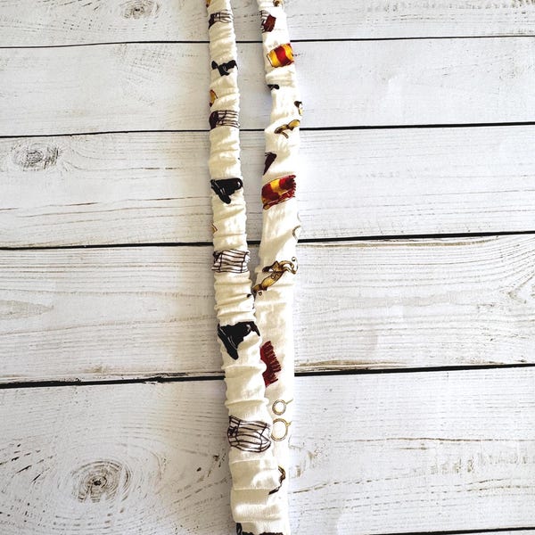 May include: A white fabric lanyard with a repeating pattern of black silhouettes, gold accents, and red and gold crests. The lanyard has a metal clasp and ring at the bottom. The fabric is gathered, creating a textured appearance.