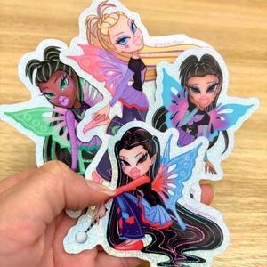 Könnte beinhalten: Eine Sammlung holografischer Aufkleber mit stilisierten Feenpuppen mit großen Augen und Lippen. Jede Puppe hat einzigartige Haare, Outfits und bunte Schmetterlingsflügel. Die Aufkleber haben ein glitzerndes, irisierendes Finish. Der Text "@HiddenPixie" ist sichtbar.