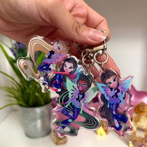 May include: Clear acrylic keychains featuring Bratz-style fairy characters with long hair, wings, and stylish outfits. The keychains have a silver metal clasp and rings for attaching to keys or bags. The characters are in shades of purple, blue, and pink.