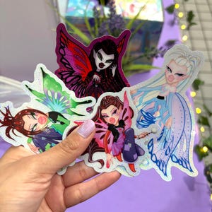 Puede incluir: Una mano que sostiene una colección de cinco pegatinas brillantes con personajes de hadas estilizados. Cada pegatina tiene un diseño único con colores vibrantes, incluyendo rojo, morado, verde y azul, y alas detalladas. Las hadas tienen rasgos exagerados.