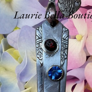 Può includere: Collana con pendente color argento con un pendente rettangolare con tre pietre preziose colorate: rosso, blu e giallo. Il pendente è decorato con dettagli floreali e un piccolo ciondolo a forma di cuore con la scritta "Made with Love".