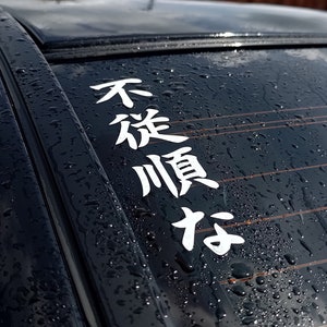 May include: A black car with raindrops on the back window. A white vinyl sticker with Japanese characters reads "不従順な" on the window.