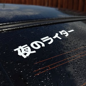 Puede incluir: Un coche negro con una pegatina blanca en la ventana trasera que dice "夜のライダー" en caracteres japoneses.