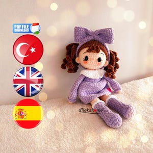 Pode incluir: Uma boneca de crochê feita à mão em posição sentada, usando um vestido lilás, botas combinando e um laço grande. A boneca tem cabelo castanho encaracolado e uma gola branca. A imagem inclui o texto "PDF FILE DOWNLOAD" e bandeiras.