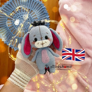 May include: A handmade crochet Eeyore plush toy in light blue, with pink inner ears and a pink belly. The plush has black eyes and a small black tuft of hair. The background includes a blue doily, fairy lights, and a pink blanket.