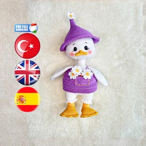 Puede incluir: Una muñeca de pato tejida a ganchillo de color blanco que lleva un sombrero morado con una pequeña flor blanca en la parte superior y un vestido de peto morado con dos flores blancas tipo margarita en el frente. El pato tiene pico y pies amarillos.
