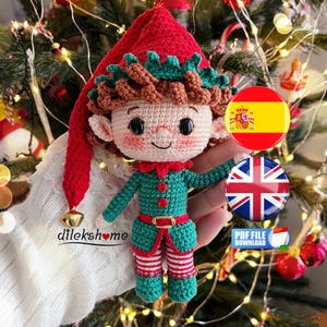 May include: A crocheted elf doll with a red hat and green and red outfit. The doll has a smiling face and brown hair. The background includes Christmas tree decorations and fairy lights. The text "dilekshome" is visible. Includes a PDF file download.