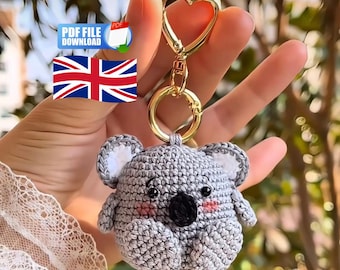 May include: A crocheted gray koala keychain with a gold-colored heart-shaped clasp. The koala has a black nose, pink cheeks, and white inner ears. The keychain is held up in front of a blurred green background.