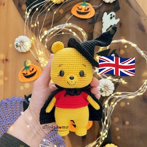 May include: A yellow crochet bear wearing a black witch hat and cape, with a red shirt. The bear is holding a UK flag and is surrounded by Halloween decorations, including pumpkins and lights. The bear's name is dilekshome.