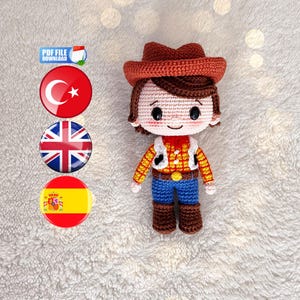 Puede incluir: Muñeco de ganchillo de un personaje de vaquero, con sombrero marrón, camisa de cuadros amarilla, vaqueros azules y botas marrones. El muñeco tiene una cara sonriente, sobre un fondo blanco.