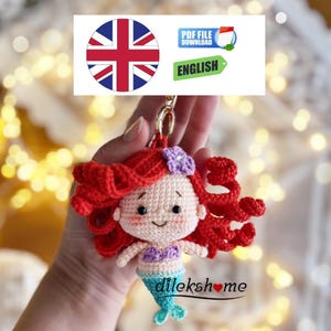 May include: A crocheted mermaid keyring with red hair, a purple flower, and a blue tail. The keyring is attached to a gold-coloured ring. The image also includes a Union Jack flag and the word "ENGLISH".