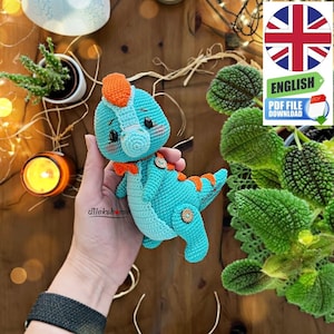 May include: A crocheted dinosaur toy with a blue body, orange accents, and black button eyes. The dinosaur is being held in a person's hand.