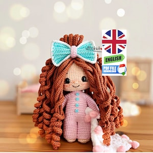 May include: Crochet amigurumi doll with brown curly hair, a blue bow, and a pink and blue pajama outfit. The doll is holding a white blanket with pink accents.