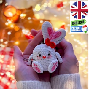 May include: A white crocheted bunny with pink ears and a red bow. The bunny has a heart-shaped nose and pink feet. The bunny is being held in someone's hands.