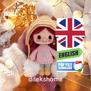 May include: Crochet doll wearing a pink sweater with a snowflake design, a yellow hat with a white pom-pom, and blue jeans. The doll has red hair and brown eyes. The doll is holding a white pom-pom in each hand. The image includes the text "ENGLISH", "PDF FILE DOWNLOAD", and "PDF".