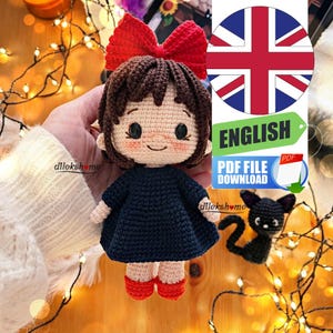 May include: Crochet amigurumi doll with brown hair, a red bow, and a black dress. The doll is wearing red shoes and has a smiling face. The doll is sitting on a wooden surface with a black cat amigurumi doll in the background.