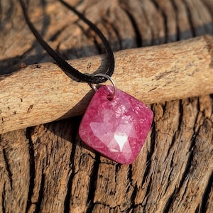Puede incluir: Un colgante de piedra rosa en un collar de cordón negro. El colgante es una piedra facetada de forma cuadrada con un acabado brillante y pulido.