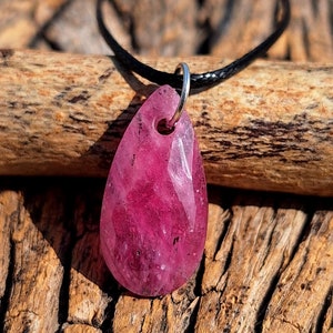 May include: A teardrop-shaped pendant necklace featuring a faceted, deep pink gemstone. The pendant is suspended from a black cord and hangs against a backdrop of textured wood. The gemstone has a polished, reflective surface.