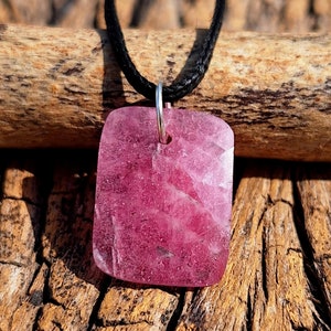 May include: A pink gemstone pendant on a black cord necklace. The pendant is a square shape with a faceted surface.