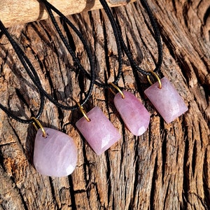 May include: Four pink gemstone pendants on black cords. The pendants are rectangular and have a polished, smooth finish. The cords are tied together at the top.