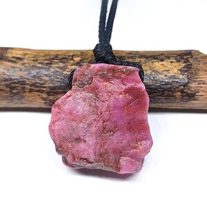 Peut inclure: Un pendentif en pierre brute de forme irrégulière, aux teintes roses et rougeâtres, suspendu à un cordon noir fin. La pierre présente une surface naturelle texturée avec des fissures visibles et des variations de couleur, nouée au cordon.