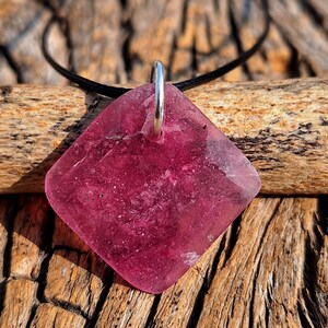 May include: A pink glass pendant with a silver bail, hanging from a black cord. The pendant is shaped like a diamond and has a sparkly, textured surface.