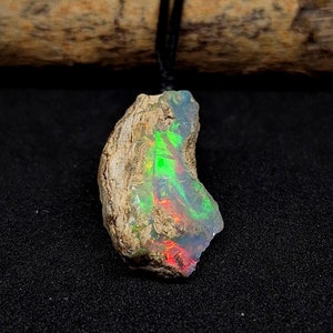 Könnte beinhalten: Ein roher, natürlicher Opal-Anhänger mit einem grün-rot-blauen, irisierenden Schimmer. Der Opal ist in einem rauen, braunen und grauen Stein gefasst. Der Anhänger hängt an einer schwarzen Schnur.