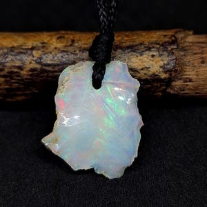 May include: A raw opal pendant with a black cord. The opal is a light, translucent color with iridescent flashes of green, pink, and blue. The stone has a rough, natural shape and is suspended from a knotted black cord.